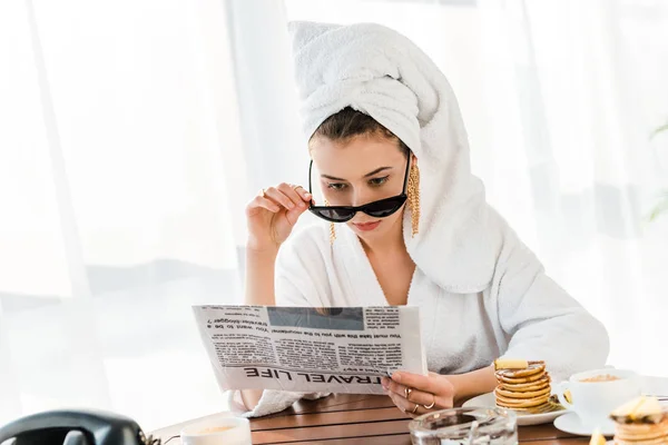 Femme élégante en peignoir, lunettes de soleil et bijoux avec serviette sur la tête lecture journal le matin — Photo de stock