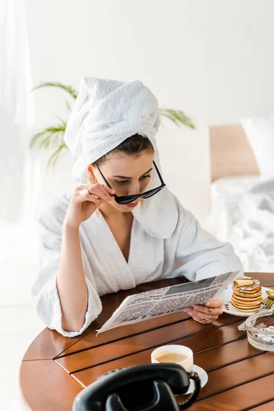 Frau im Bademantel, Sonnenbrille und Schmuck mit Handtuch auf dem Kopf liest morgens Zeitung — Stockfoto