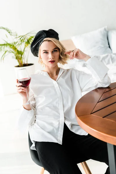 Elegante mujer rubia en boina negra y camisa blanca sentada a la mesa y sosteniendo vaso con vino tinto - foto de stock