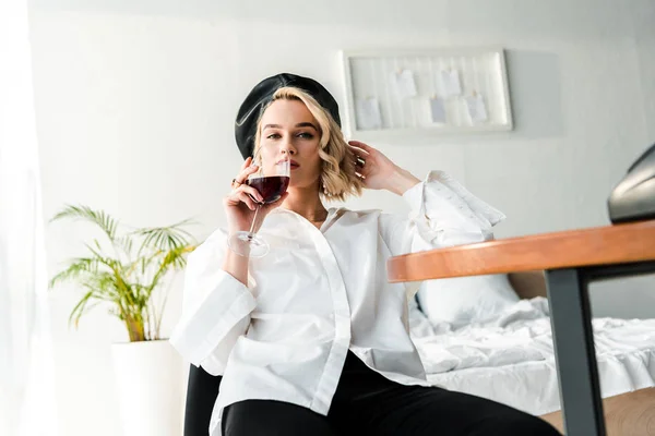 Elegante mujer rubia en boina negra y camisa blanca sentada a la mesa y bebiendo vino tinto - foto de stock