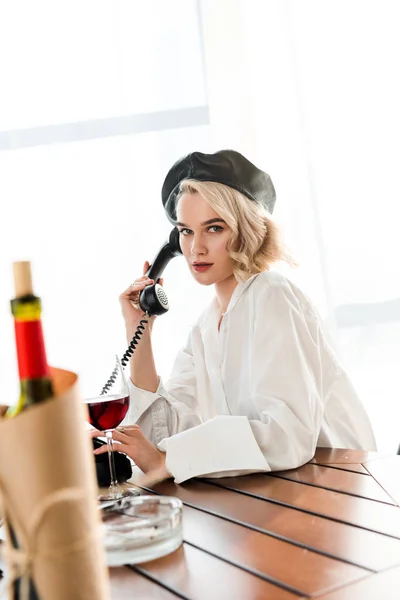 Enfoque selectivo de mujer rubia elegante en boina negra y camisa blanca sentado en la mesa y hablando o teléfono retro - foto de stock