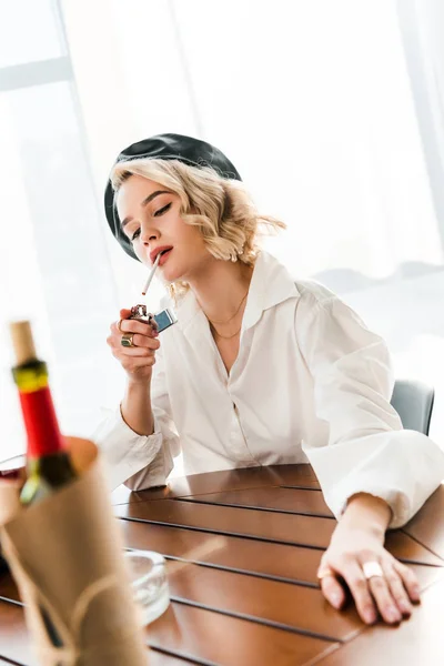 Selective focus of elegant blonde woman in black beret and white shirt lighting up cigarette near wine bottle — Stock Photo