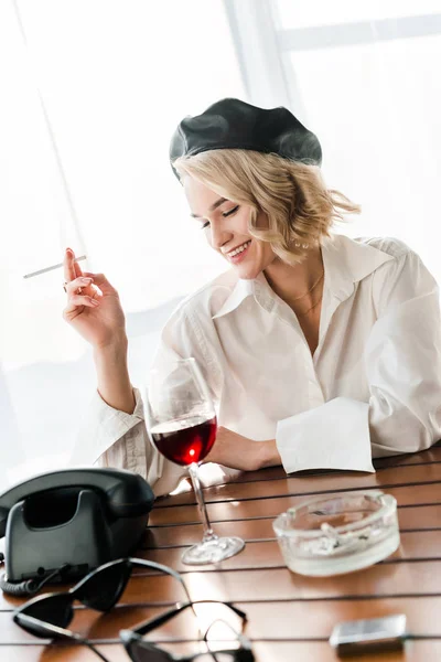 Elegant happy blonde woman in black beret and white shirt smoking cigarette near glass with red wine and retro telephone — Stock Photo