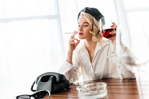 Elegant dreamy blonde woman in black beret and white shirt with closed eyes smoking cigarette and holding glass with red wine — Stock Photo