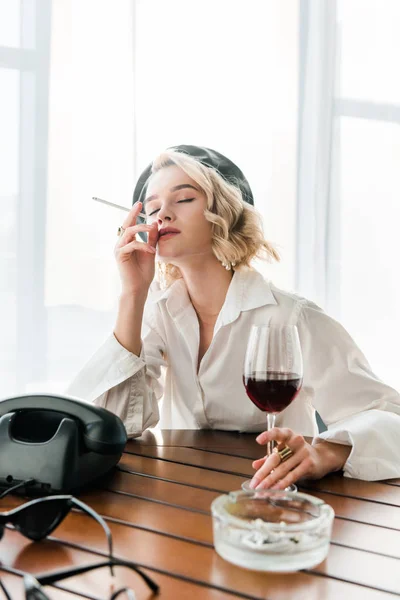 Élégante femme blonde rêveuse en béret noir et chemise blanche tenant cigarette et verre avec vin rouge — Photo de stock