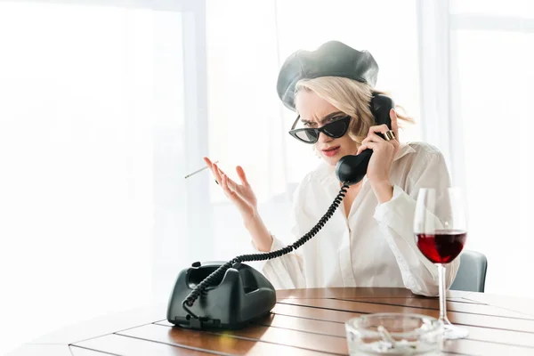 Élégante femme blonde insatisfaite en béret noir et lunettes de soleil fumer cigarette tout en parlant sur téléphone rétro près d'un verre de vin rouge — Photo de stock