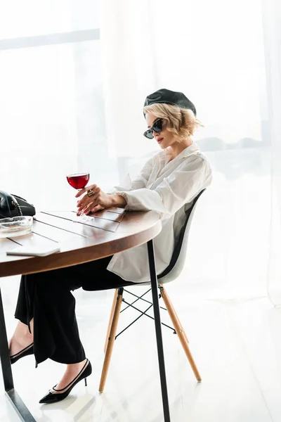 Elegant blonde woman in black beret and sunglasses sitting at table with glass of red wine — Stock Photo