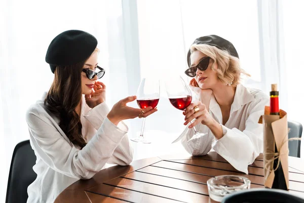 Elegante morena y mujeres rubias en boinas negras y gafas de sol tintineando con copas de vino tinto en la mesa de madera — Stock Photo