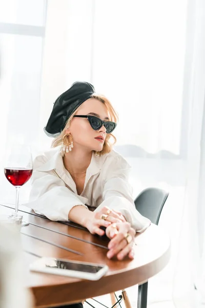 Élégante femme blonde rêveuse en béret noir et lunettes de soleil assises à table avec verre de vin rouge près du smartphone — Photo de stock