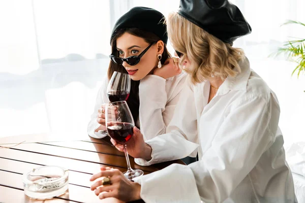 Elegant brunette and blonde women in black berets and sunglasses drinking red wine at wooden table — Stock Photo