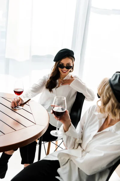 Vista aérea de elegante mujer morena sonriente en boina negra y gafas de sol bebiendo vino tinto en mesa de madera con amigo rubio - foto de stock