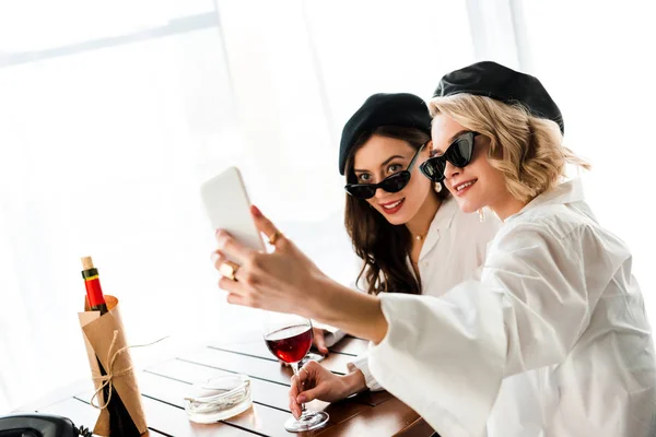 Elegante morena sonriente y mujeres rubias en boinas negras y gafas de sol bebiendo vino tinto y tomando selfie - foto de stock