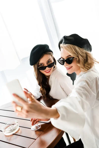 Selective focus of elegant smiling brunette and blonde women in black berets and sunglasses taking selfie on smartphone — Stock Photo