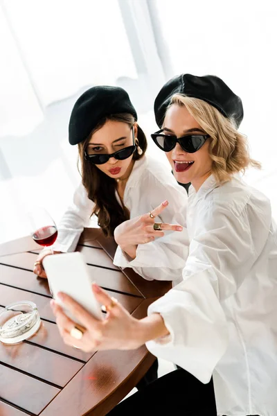Mulheres morenas e loiras elegantes em boinas pretas e óculos de sol bebendo vinho tinto e tirando selfie enquanto amuam os lábios e mostram sinal de vitória — Fotografia de Stock
