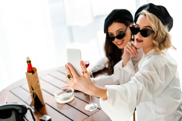 Selective focus of elegant brunette and blonde women in black berets and sunglasses drinking red wine and taking selfie — Stock Photo