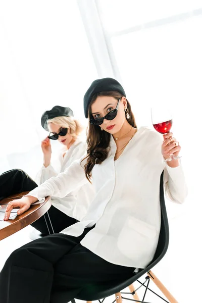 Selective focus of elegant brunette woman in black beret and sunglasses with glass of red wine sitting near blonde friend — Stock Photo