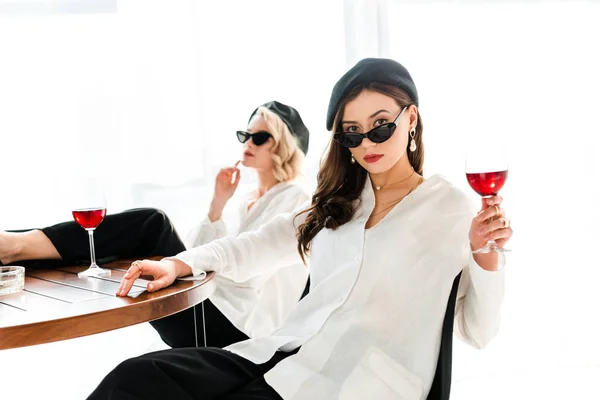 Foyer sélectif de femme brune élégante en béret noir et lunettes de soleil avec verre de vin rouge assis près d'un ami — Photo de stock