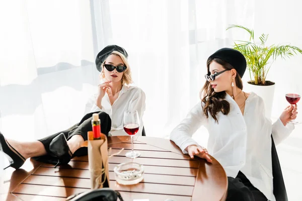 Mulheres morenas e loiras elegantes em boinas pretas e óculos de sol bebendo vinho tinto enquanto se sentam à mesa — Fotografia de Stock