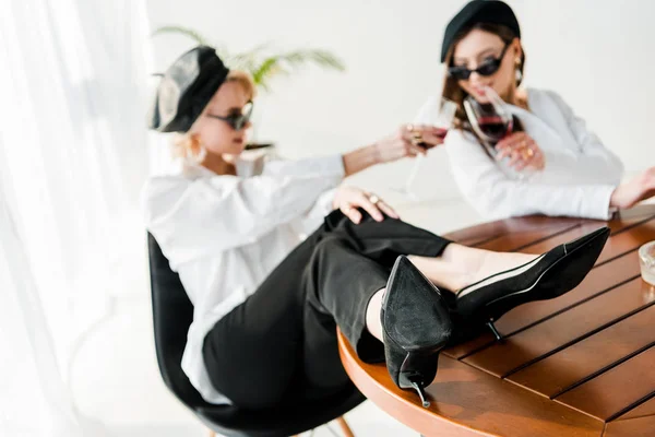 Selektiver Fokus der eleganten Frau in schwarzer Baskenmütze und Sonnenbrille mit Beinen auf dem Tisch, die mit Freund klirrt — Stockfoto