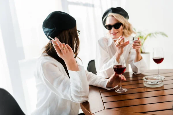 Morena y mujeres rubias en boinas negras y gafas de sol bebiendo vino tinto, hablando y fumando cigarrillos - foto de stock