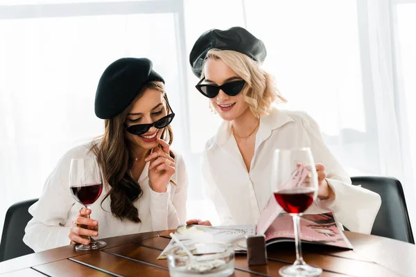 Брюнетка и блондинка, улыбающиеся женщины в черных беретах и солнечных очках, пьющие красное вино и читающие журналы — стоковое фото