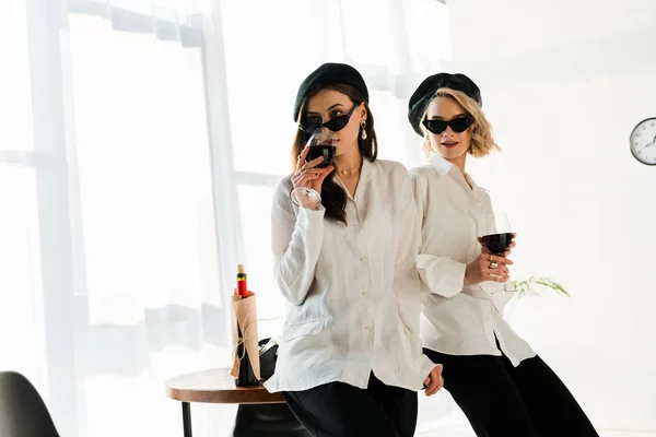 Élégante brune et blonde aux bérets noirs et lunettes de soleil buvant du vin rouge — Photo de stock