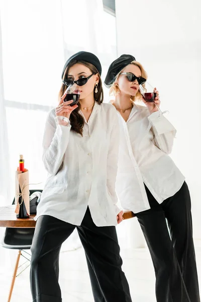 Élégante brune et amies blondes aux bérets noirs et lunettes de soleil buvant du vin rouge — Photo de stock