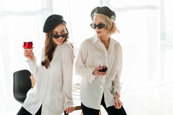 Élégante brune souriante et amies blondes aux bérets noirs et lunettes de soleil buvant du vin rouge — Photo de stock