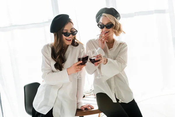 Elegant happy brunette and blonde women in black berets and sunglasses laughing, smoking and drinking red wine — Stock Photo