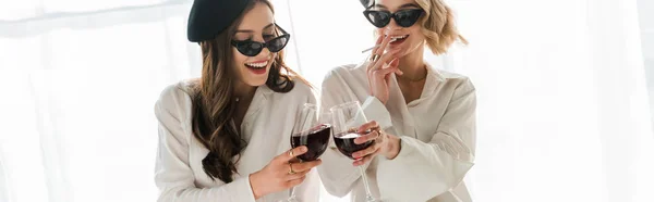 Plan panoramique d'élégantes femmes brunes et blondes heureuses en bérets noirs et lunettes de soleil riant, fumant et buvant du vin rouge — Photo de stock