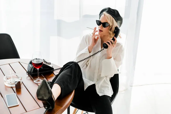 Mulher loira elegante em boina preta e óculos de sol sentado com a perna na mesa, fumando e falando telefone retro — Fotografia de Stock