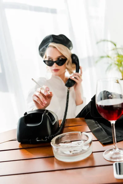 Foco seletivo da mulher loira elegante em boina preta e óculos de sol fumando e falando telefone retro perto de vidro com vinho tinto — Fotografia de Stock