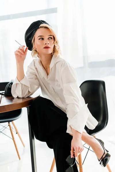 Elegante mujer rubia en boina negra sentada en silla en pose y fumando cigarrillo - foto de stock