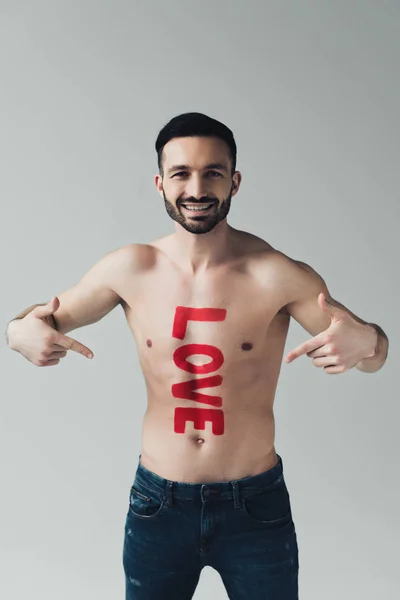 Vista frontal del hombre sin camisa sonriente señalando con los dedos la inscripción en el cuerpo aislado en gris - foto de stock