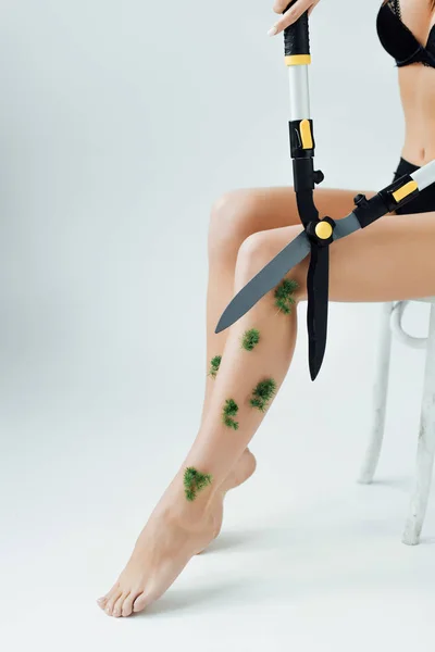 Cropped view of woman sitting on chair and cutting plants on legs with big scissors on grey — Stock Photo