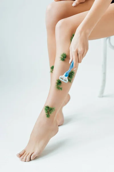 Cropped view of woman sitting on chair and shaving plants on legs on grey — Stock Photo