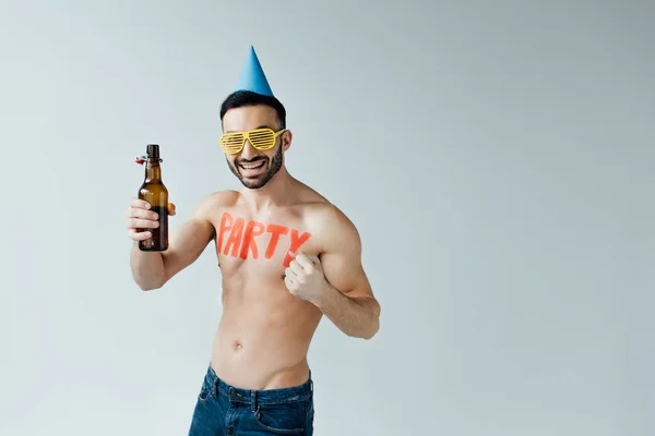 Homem sorridente de óculos e boné de festa segurando cerveja e olhando para a câmera isolada em cinza — Fotografia de Stock