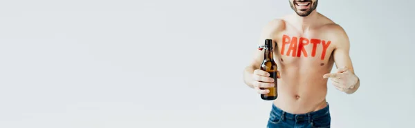 Panoramic shot of smiling bearded man pointing with finger at beer bottle isolated on grey — Stock Photo