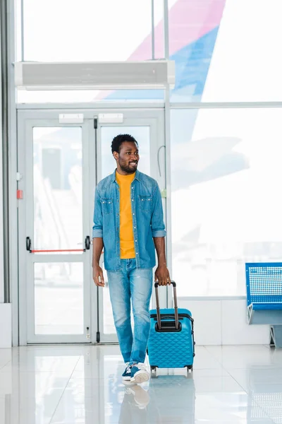 Sonriente hombre afroamericano feliz caminando con maleta a lo largo de la sala de espera en el aeropuerto - foto de stock