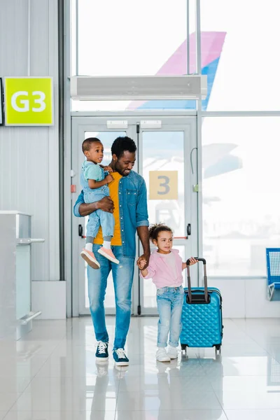 Улыбающийся африканский отец-американец, идущий с детьми и багажом вдоль зала ожидания в аэропорту — стоковое фото