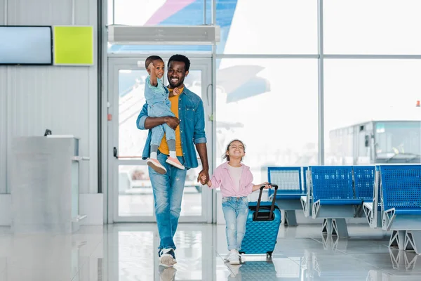 Lächelnder afrikanisch-amerikanischer Vater läuft mit glücklichen Kindern und Gepäck entlang der Wartehalle im Flughafen — Stockfoto