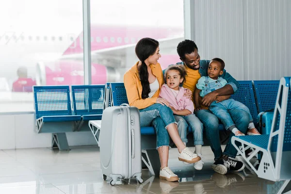 Улыбающаяся африканская американская семья с багажом и детьми, сидящими в аэропорту — стоковое фото