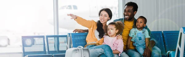 Colpo panoramico di sorridente famiglia afroamericana con bagagli e bambini seduti in aeroporto mentre la madre punta con il dito lontano — Foto stock