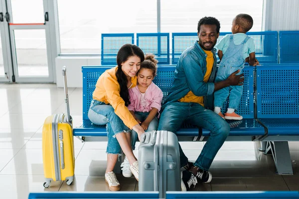 Alegre familia afroamericana con bolsas de viaje y niños sentados en el aeropuerto - foto de stock