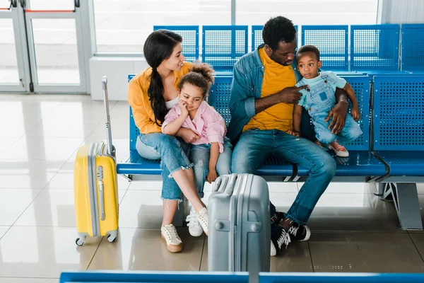 Alegre familia afroamericana con bolsas de viaje y niños sentados en el aeropuerto en la sala de espera - foto de stock