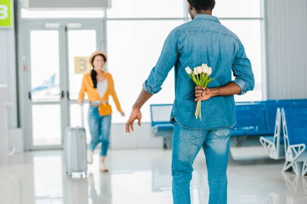 Selektiver Fokus eines afrikanisch-amerikanischen Mannes, der mit einem Koffer auf seine glückliche Freundin zugeht, während er im Flughafen Tulpen hinter seinem Rücken versteckt — Stockfoto