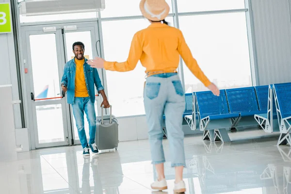 Foco seletivo de homem americano africano feliz com bagagem andando em direção a namorada no aeroporto — Fotografia de Stock