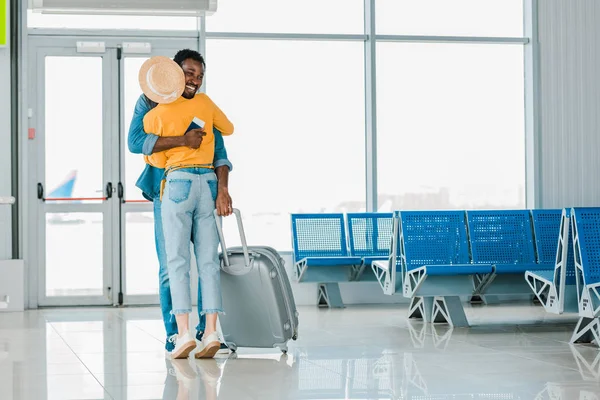 Hombre afroamericano feliz con bolsa de viaje abrazando novia en aeropuerto - foto de stock