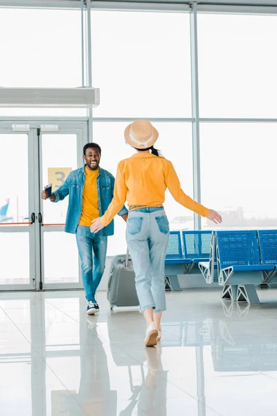 Hombre afroamericano feliz con la maleta caminando hacia la novia en el aeropuerto - foto de stock