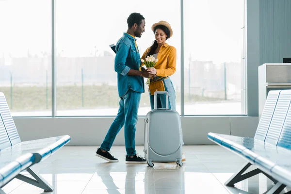 Smiling african american man gifting bouquet of tulips to girlfriend with suitcase in airport — Stock Photo